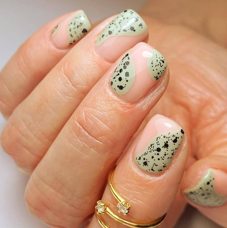 A closeup of a woman's fingernails with a glossy nude nail polish that has a combination of an abstract design in sage green and black polish to add dots and speckles 