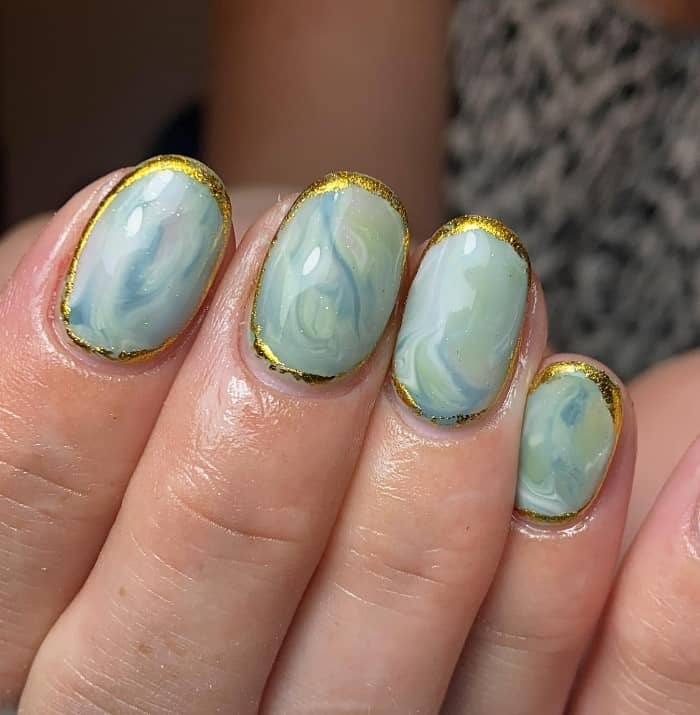 A closeup of a woman's fingernails with sage green and blue nail polish swirl together that has a loose gold outline