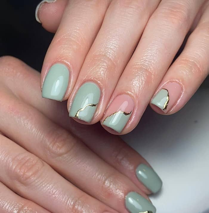 A closeup of a woman's fingernails with a combination of a nude and sage green nail polish that has jagged gold lines on select nails