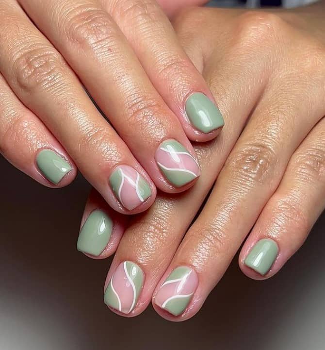 A closeup of a woman's fingernails with a combination of pale pink and sage green nail polish that has swirl patterns in white