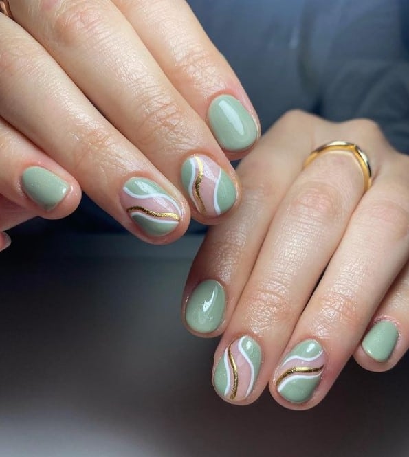 A closeup of a woman's fingernails with a combination of nude and sage green nail polish that has gold and white accents on select nails