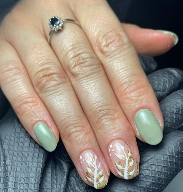 A closeup of a woman's fingernails with a combination of nude and sage green nail polish that has white-and-gold seaweed nail art