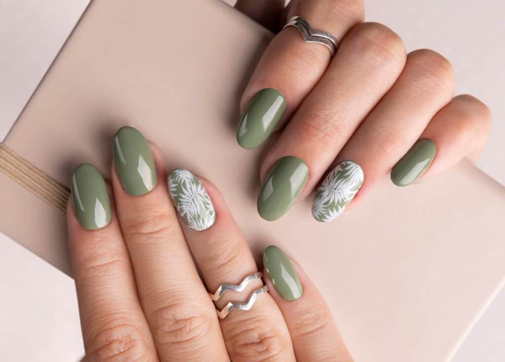 A woman's fingernails with a glossy sage green nail polish that has bright white tropical-leaf nail designs on select nails