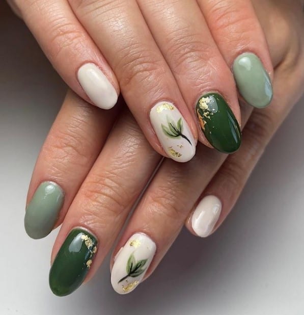 A closeup of a woman's fingernails with a combination of white and different shades of green nail polish that has gold foil flakes and classy leaf nail art 