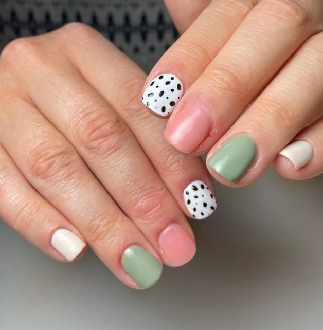 A closeup of a woman's fingernails with a combination of pastel pink, sage green, and white nail polish that has polka dots on white nails