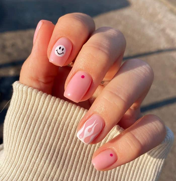 A closeup of a woman's fingernails with baby pink nail polish base that has a smiley face, white fire and red dots nail designs