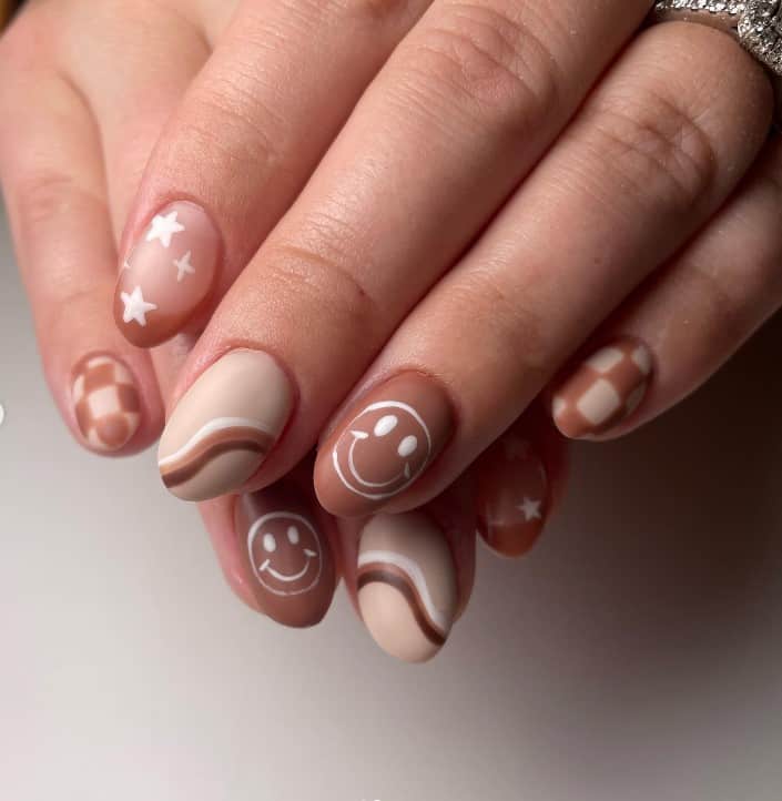 A closeup of a woman's fingernails with mocha shades nail polish that has smiley face, checker patterns, stars, and swirls nail art designs
