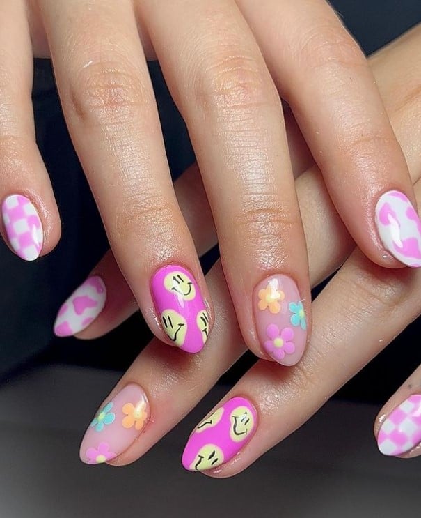 A closeup of a woman's fingernails with fuchsia nail polish that has pale yellow smiley face accents, pastel flowers, hearts and checkers on white polish 