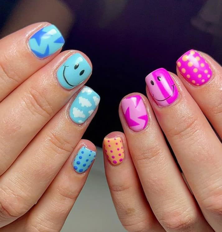 A closeup of a woman's fingernails with blue and purple nail polish that has a smiley face, dots and clouds nail designs