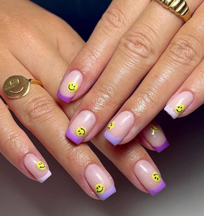 A closeup of a woman's fingernails with a nude nail polish base that has French tips in different shades of purple and a classic yellow smiley face nail design