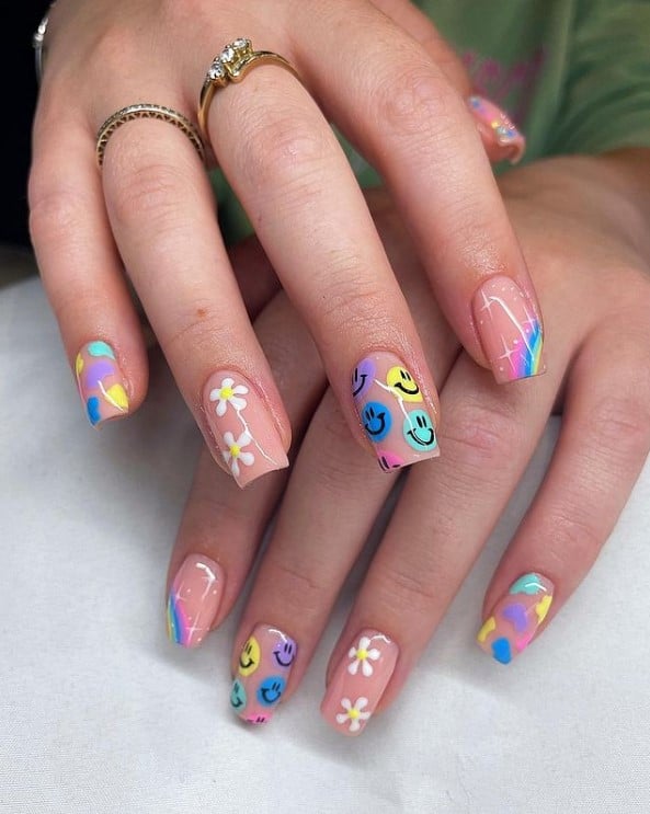 A woman's fingernails with a glossy nude nail polish that has rainbows, flowers, smiley faces and light flares nail designs