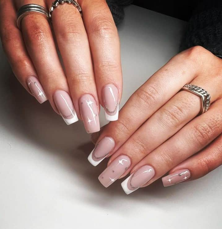 A woman's hands with a nude pink nail polish base that has white French tips on select nails, white shining stars and curved lines in silver glitter