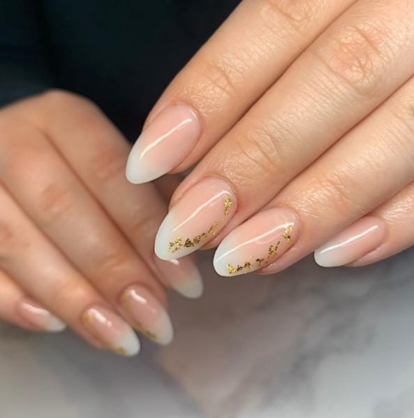 A closeup of a woman's fingernails with beige ombré nails that has gold flakes on select nails 