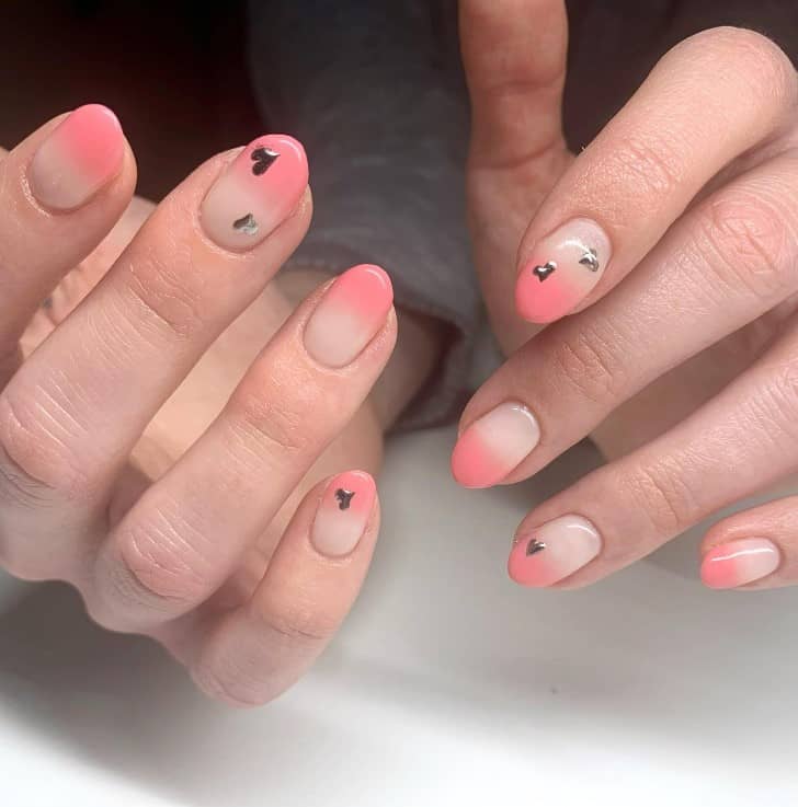 A closeup of a woman's fingernails with a beige nail polish that has peach tips and tiny metallic heart details on select nails 