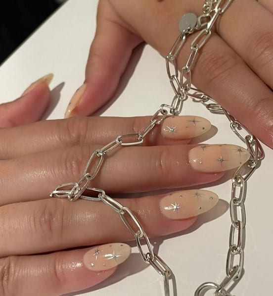 A closeup of a woman's fingernails with almond beige nails that has stars in metallic silver polish