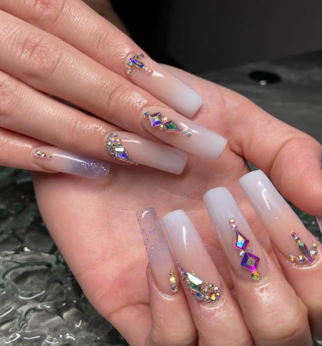 A closeup of a woman's fingernails with beige ombré nails that has crystals in various shapes and cool colors