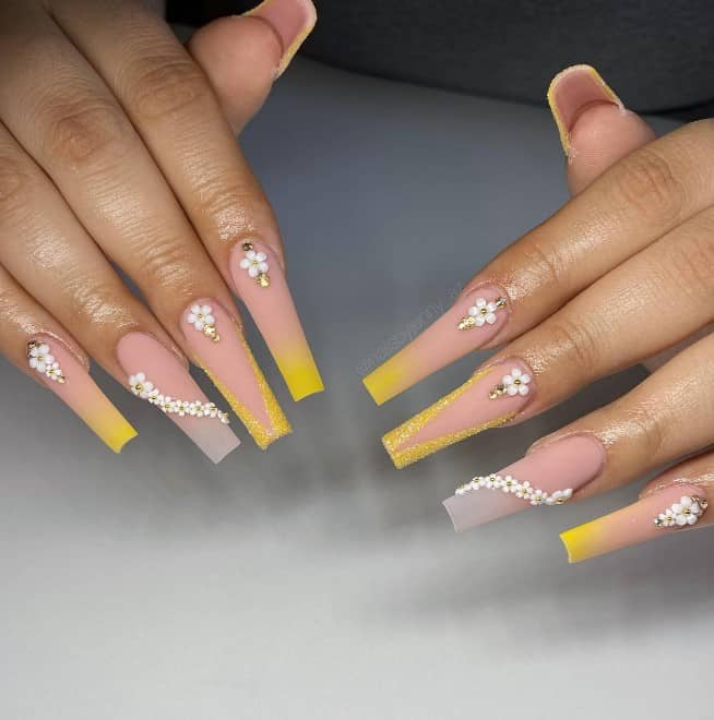 A woman's fingernails with matte nude ombré nails that has yellow ombré,  textured tips and white 3D flower stickers
