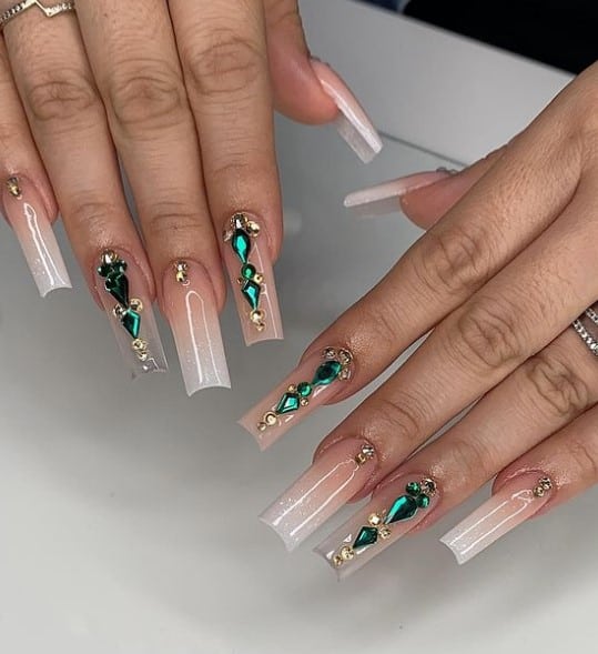 A woman's fingernails with a nude nail polish that has a shimmery and jelly finish and emerald crystals