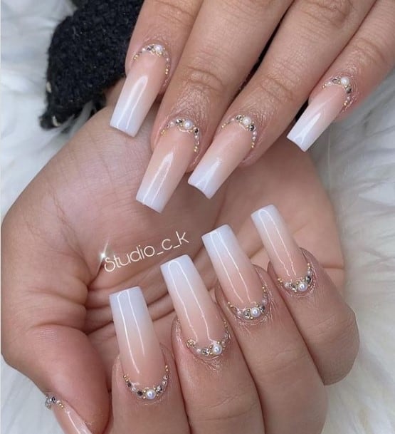 A closeup of a woman's long coffin fingernails with beige ombre nail polish that has tiny rhinestones and pearls