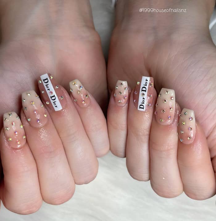 A closeup of a woman's fingernails with a nude manicure that has a Dior logo on select nails and gems