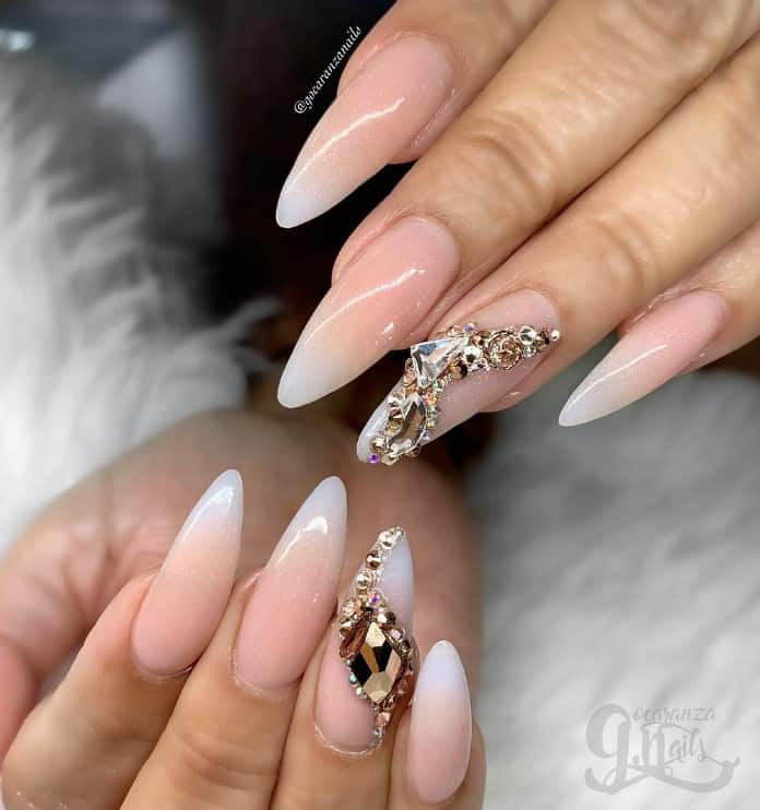A closeup of a woman's fingernails with shimmery pinkish beige-and-white ombré nails that has humongous crystals swirling on top on select nails