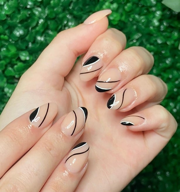 A closeup of a woman's simple and sweet fingernails with unique nude nails that has thin and thick black swirls outlined with sophisticated thin white lines