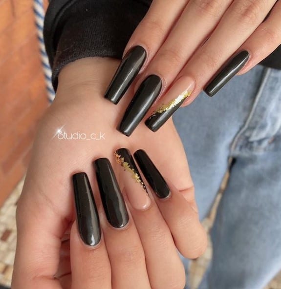 A woman's coffin fingernails with a combination of black and nude nail polish base that has streaks of glittery gold to the accent nails