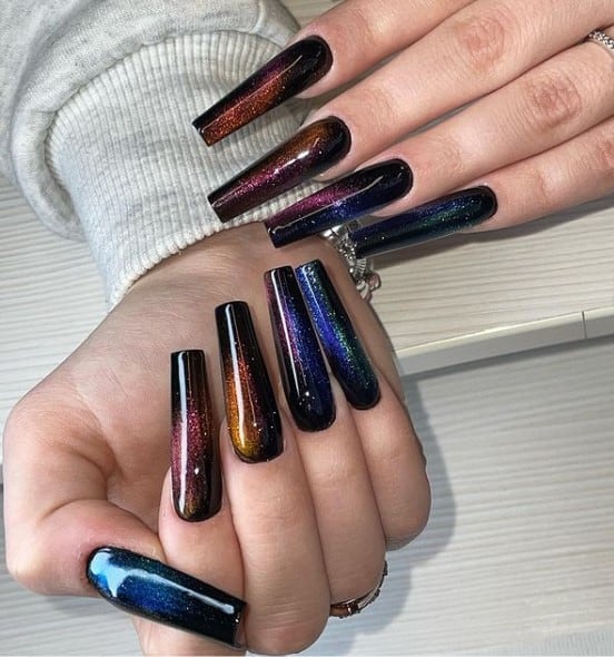 A closeup of a woman's coffin fingernails with black nail polish that has colorful glitter in a dramatic galaxy-themed design