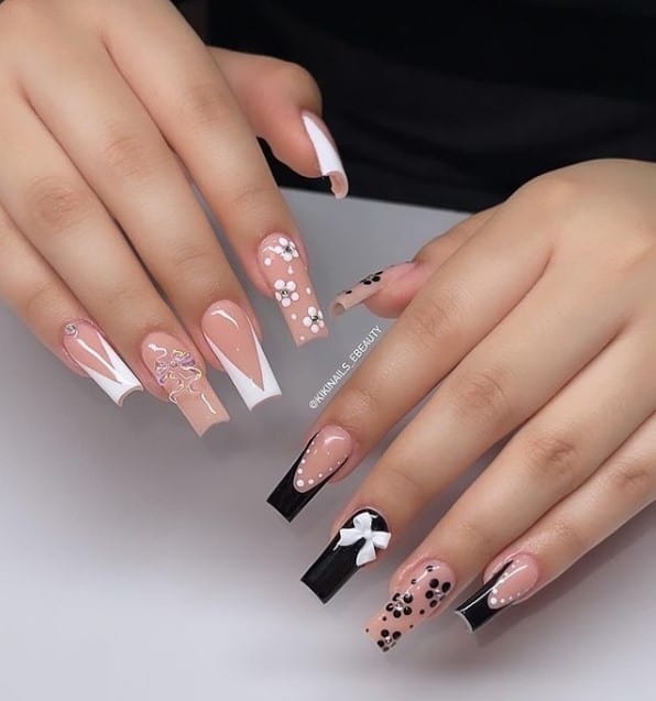 A woman's fingernails with a combination of nude and black nail polish that has 3D ribbons, black French tips with dotted outlines, handpainted florals, and V-tips