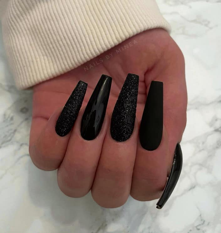 A closeup of a woman's fingernails with a combination of matte and glossy black nail polish that has black glitter on select nails 