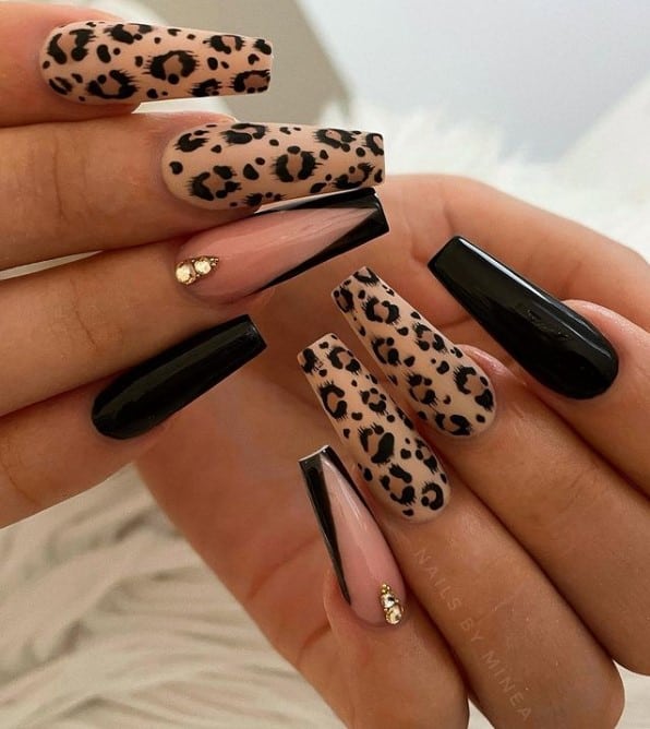 A closeup of a woman's coffin fingernails with a nude and black nail polish that has leopard print nail art and gems on select nails 