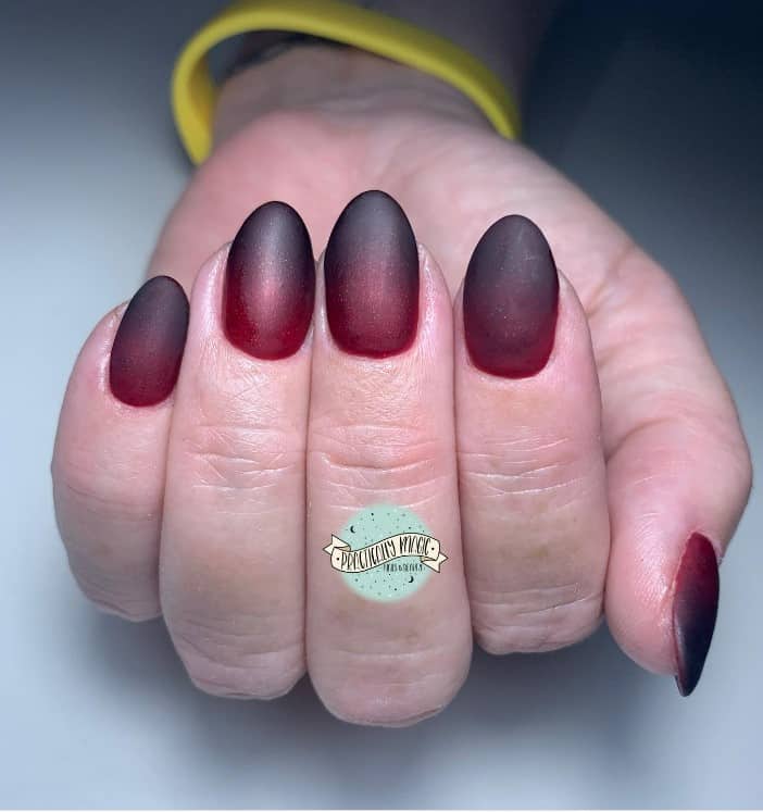 A closeup of a woman's oval shaped fingernails with a red matte to black ombré manicure 