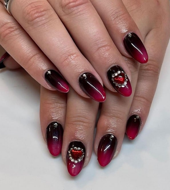 A closeup of a woman's fingernails with a glossy black-and-red ombré nails that has rhinestones and heart gems on select nails 