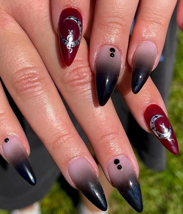 A closeup of a woman's fingernails with black ombré nails that has black-to-nude gradient paired with a blood-red accent nail embellished with a unique cat and moon design in silver