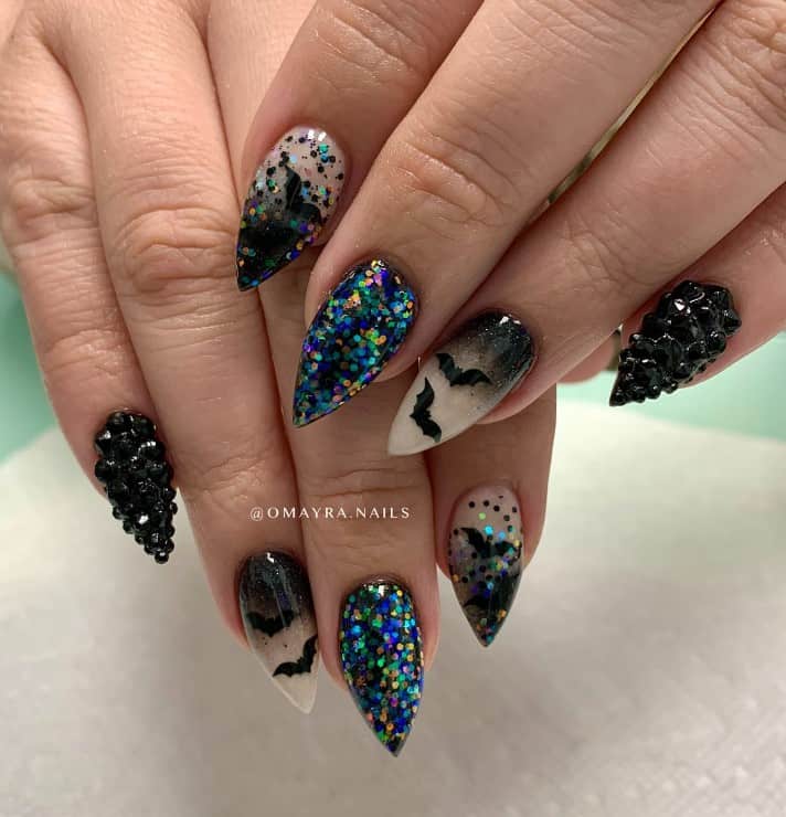 A closeup of a woman's fingernails with black ombré glitter nails with bats that has 3D black gems and multicolored glitter flakes