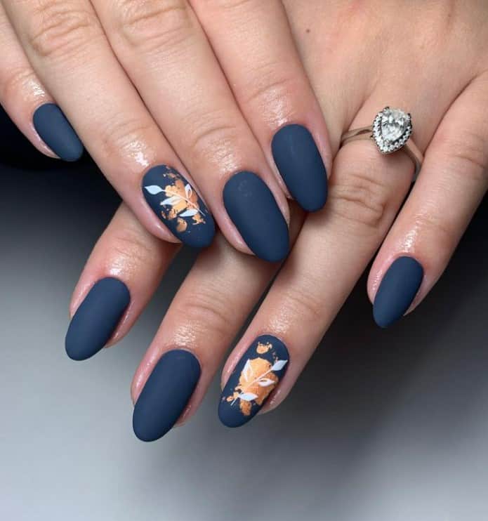 A closeup of a woman's fingernails with enchanting matte blue and gold nail design that has gold splatters and white leaves and stems at the center