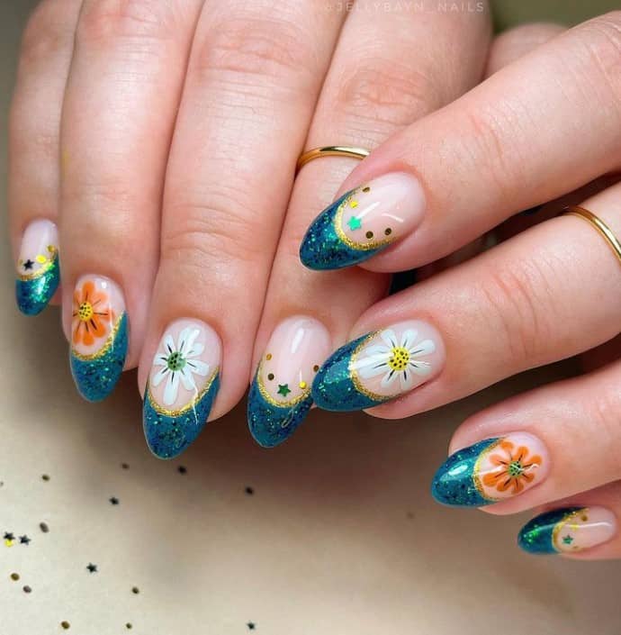 A closeup of a woman's fingernails with a nude nail polish base that has glittery teal French tips lined with gold at the bottom, dots and star sequins or intricately hand-painted flowers