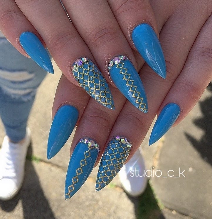 A closeup of a woman's stiletto-shape fingernails with a shade of blue nail polish that has tiny gems near the cuticles and decorated with a net-like pattern in radiant gold at the center