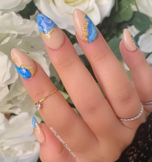A closeup of a woman's fingernails with a nude nail polish base that has two tones of blue on different sides and gold foil flakes