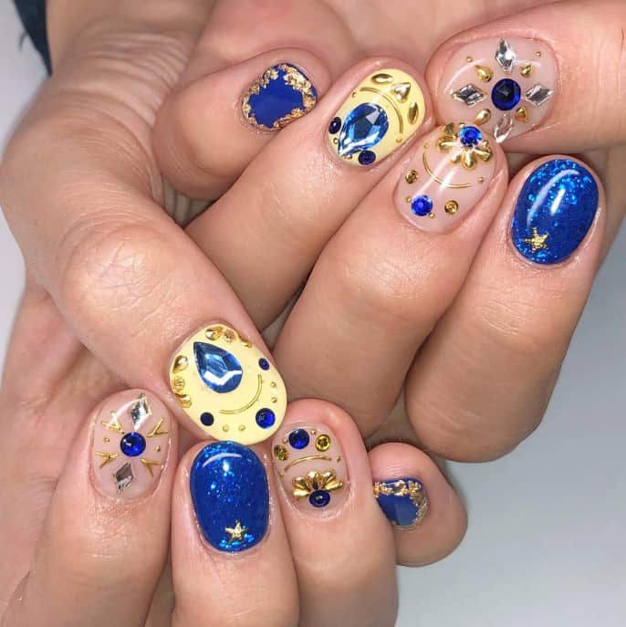 A  closeup of a woman's fingernails with a mix of sparkly blue, yellow, and nude peach nail polish base that has blue, silver, and gold gems and flecks of gold foil