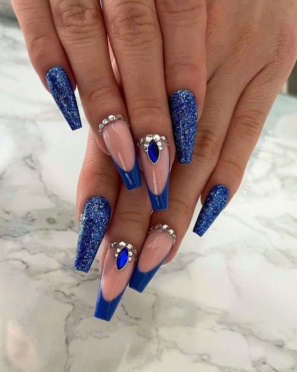 A closeup of a woman's fingernails with blue nail polish that has glitter and rhinestones under the dark blue French tips