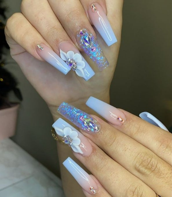 A woman's fingernails with a blue-and-nude ombré nail polish that has crystallized clear nails and 3D flower nail art