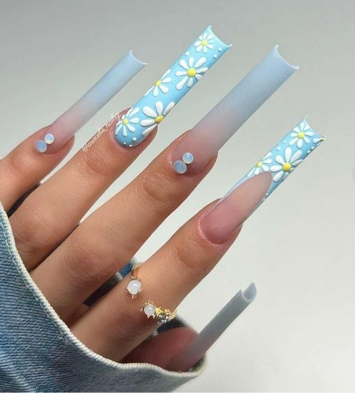A closeup of a woman's super long square fingernails with a matte French blue ombré nail polish that has 3D daisies on select nails 