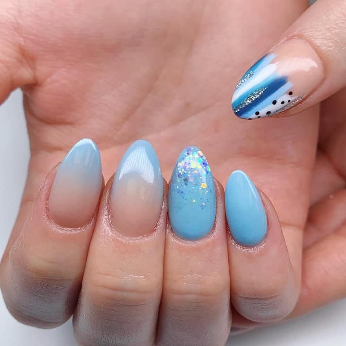 A closeup of a woman's fingernails with solid blue nail polish that has multipatterned tips and ombré glitter