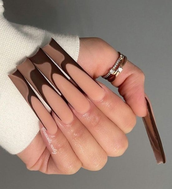 A closeup of a woman's long square fingernails with a light brown nail polish base that has thick, swoopy swirls in two shades of brown