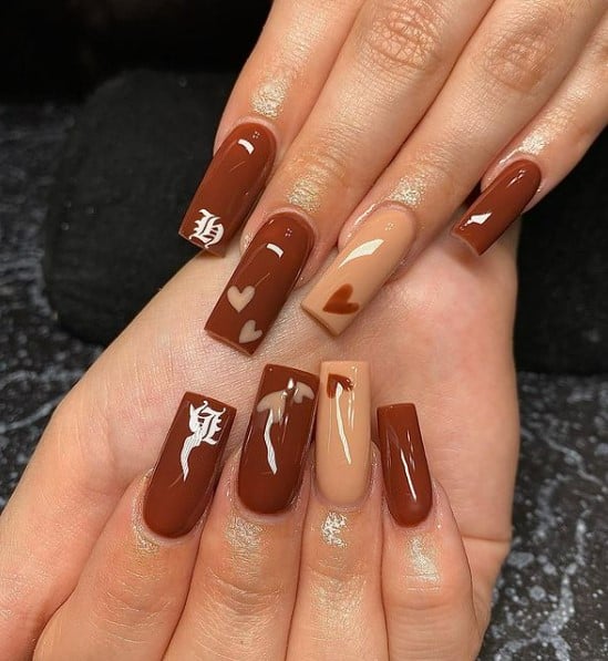 A closeup of a woman's fingernails with a combination of gingerbread brown and sand-brown color that has tiny hearts near the tips and calligraphy of initials on one nail