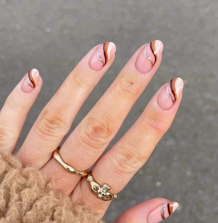 A woman's fingernails with a nude nail polish base that has brown and white swirls near the tips and a silver glitter outline