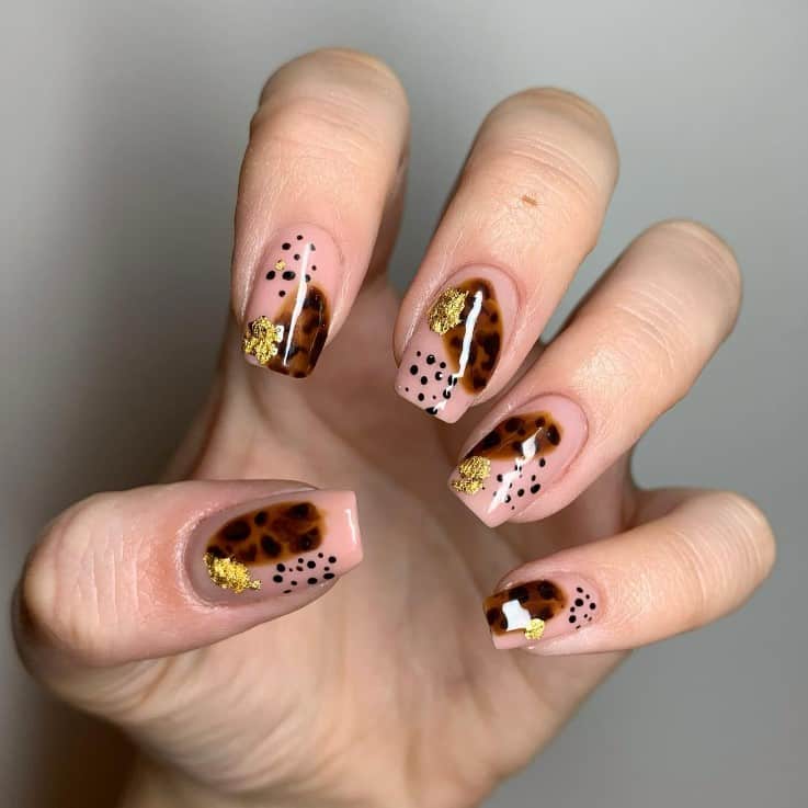 A closeup of a woman's fingernails with a nude nail polish that has speckles of leopard prints, gold flakes, and brown dots