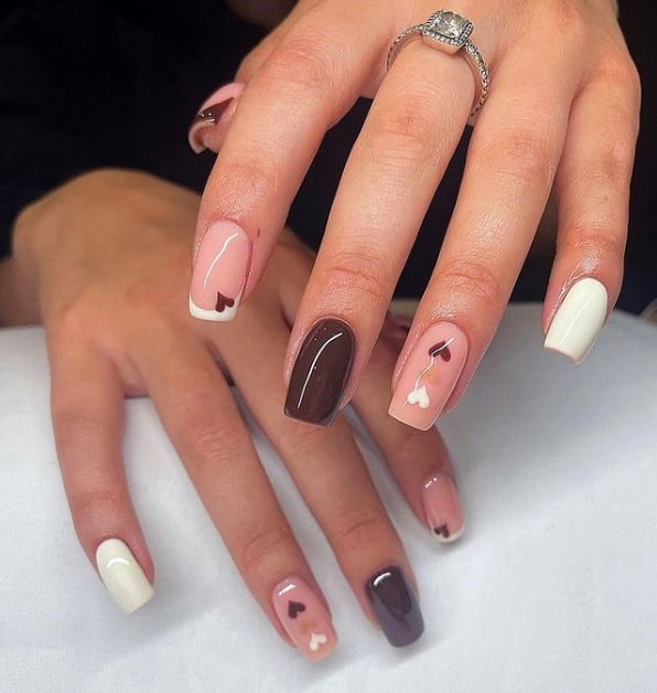A woman's fingernails with a combination of white, brown and nude nail polish that has white and brown French tips and tiny hearts on the nude accent nails