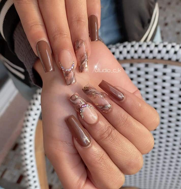 A woman's fingernails with a combination of brown and nude nail polish that has marble designs, rhinestones, and gold flakes nail designs